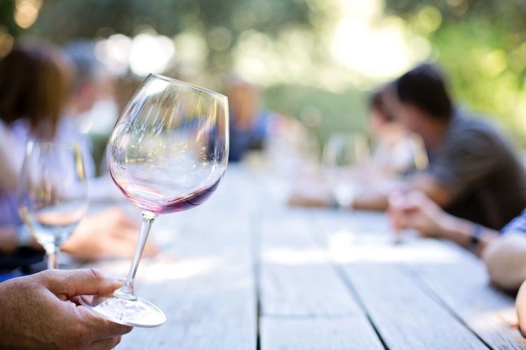 A wine glass being held in the foreground with people sitting at a long table outside in the background